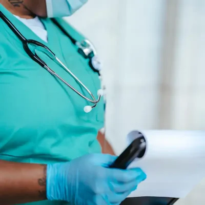 A Black woman in blue scrubs with a mask and stethoscope reads a medical chart.