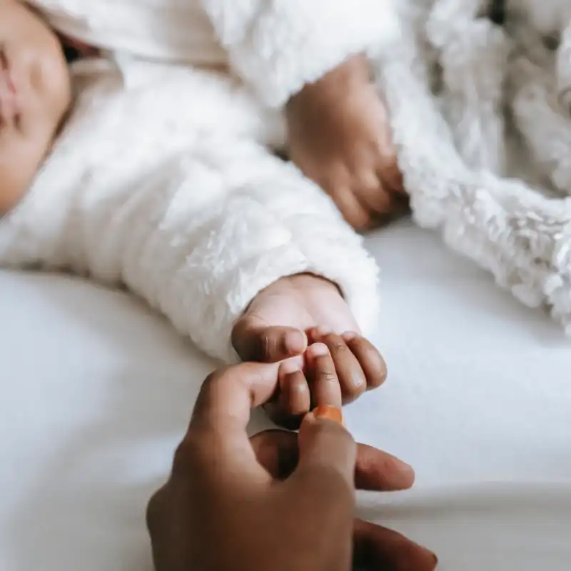 A baby wearing a fuzzy white jacket is holding a person of color's finger. 