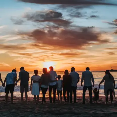 A large group of adults and children have their backs to the camera watching the sunset on a beach.