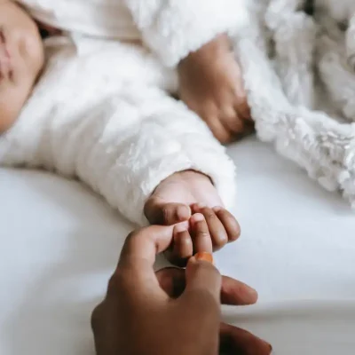 A baby wearing a fuzzy white sweater is laying down and holds the finger of an adult.