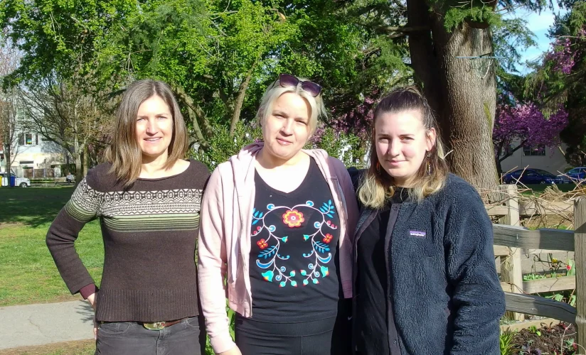 Three women are smiling and standing outside a community garden.