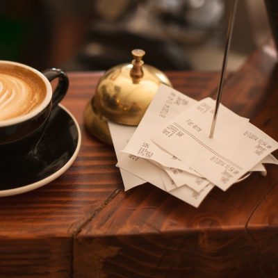 A latte is on a wooden shelf next to a bell and receipt holder spike with bills on it.