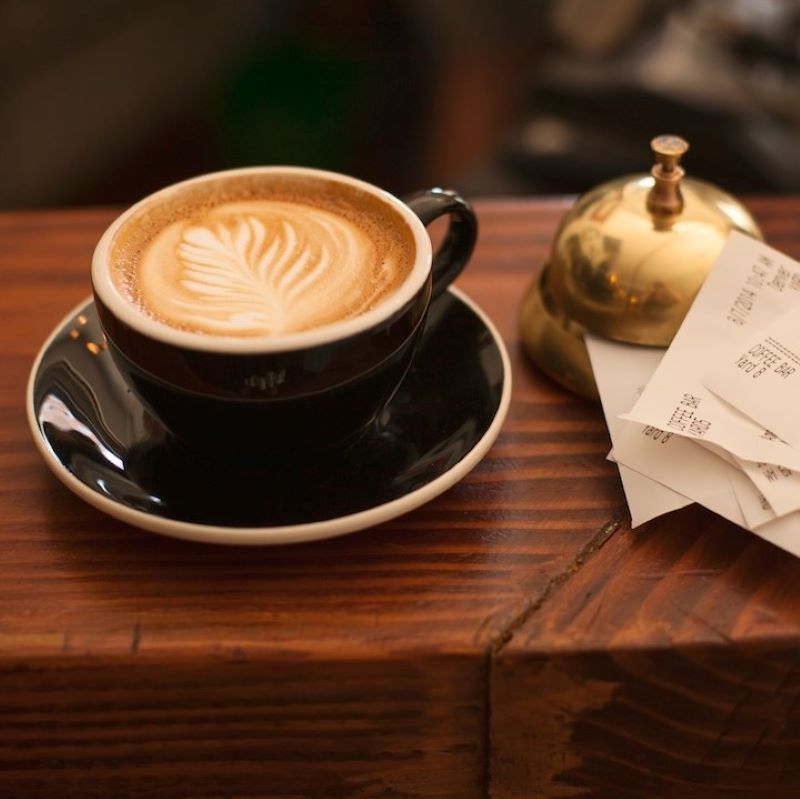 A latte sits on a wooden bar top next to a service bell and receipt holder. 