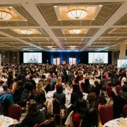 A crowded ballroom of guests sitting around tables and looking towards a stage.