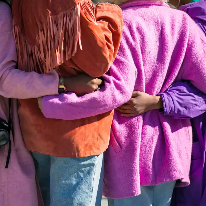 Four people with their backs to the camera have their arms around one another. Their jackets are purple, pink, or orange.