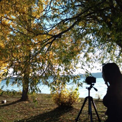 A person is standing behind a tripod, filming towards trees and a lake.