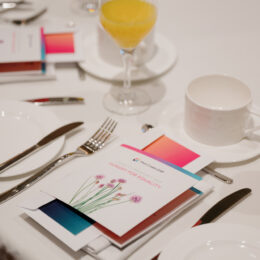 A breakfast table setting on a white tablecloth with a pink and blue program in the middle.