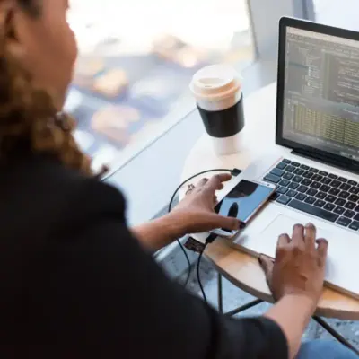 A person is typing on a laptop and reaching for their phone.