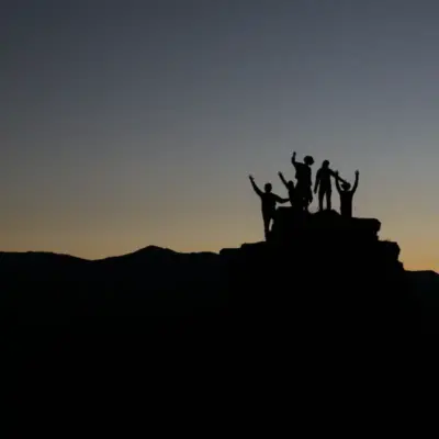 A group of people are standing on a rock with their arms raised watching the sunset.
