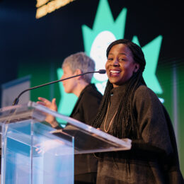 Sandy Hudson is speaking at the podium, with the ASL interpreter standing behind her.