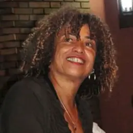 Angela Davis, a Black woman, is sitting in front of a brick wall and smiling. She has long curly hair and is wearing red lipstick.