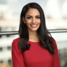 Anita Bathe is wearing a red shirt and smiling. She has long dark hair.