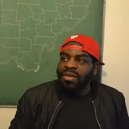 Hanif Abdurraqib, a Black man, stands in front of a green map. He is wearing a black jacket and a backwards red hat.