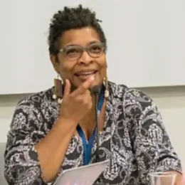 Nalo Hopkinson, a Black woman, is presenting behind a microphone. Her hair is short and she's wearing glasses and a patterned shirt.
