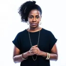 Sandy Hudson, a Black woman, is wearing a black shirt and gold jewelry, with her hands clasped in front of her.