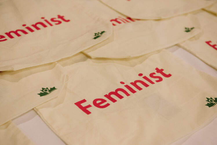 Canvas tote bags on a table that say "Feminist" in pink letters.