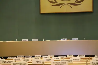An empty United Nations meeting room with rows of chairs and tables, each labeled with a country and facing a table where the UN committees sit.