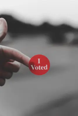 A hand holding a red sticker on their finger tip that says "I Voted"
