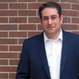 Seth Klein, BC Director of the Canadian Centre for Policy Alternatives, is wearing a blue suit and white shirt, standing in front of a brick wall.