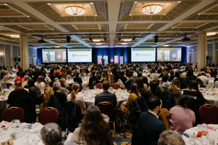 A wide shot of the full ballroom at the 2024 Equality Breakfast. Pam Palmater is delivering her keynote address from the stage, flanked by four large screens.