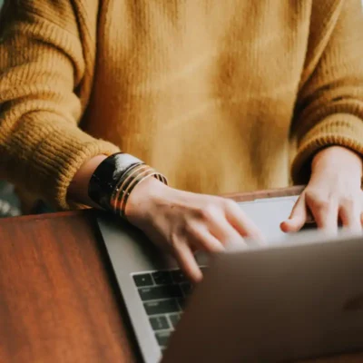 A person in a yellow sweater and bracelets is sitting at a wood table, typing on a laptop.