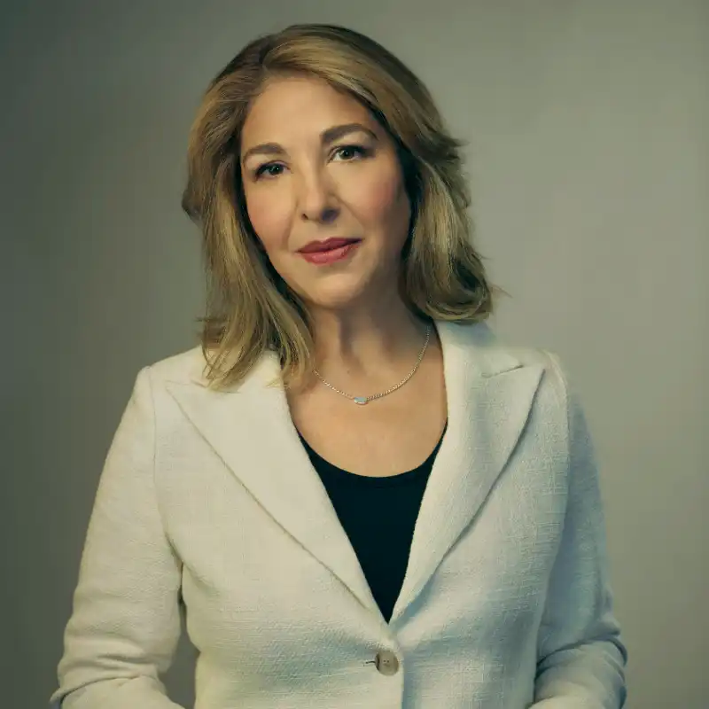 Naomi Klein is wearing a cream blazer and black shirt while smiling softly. The background is grey.